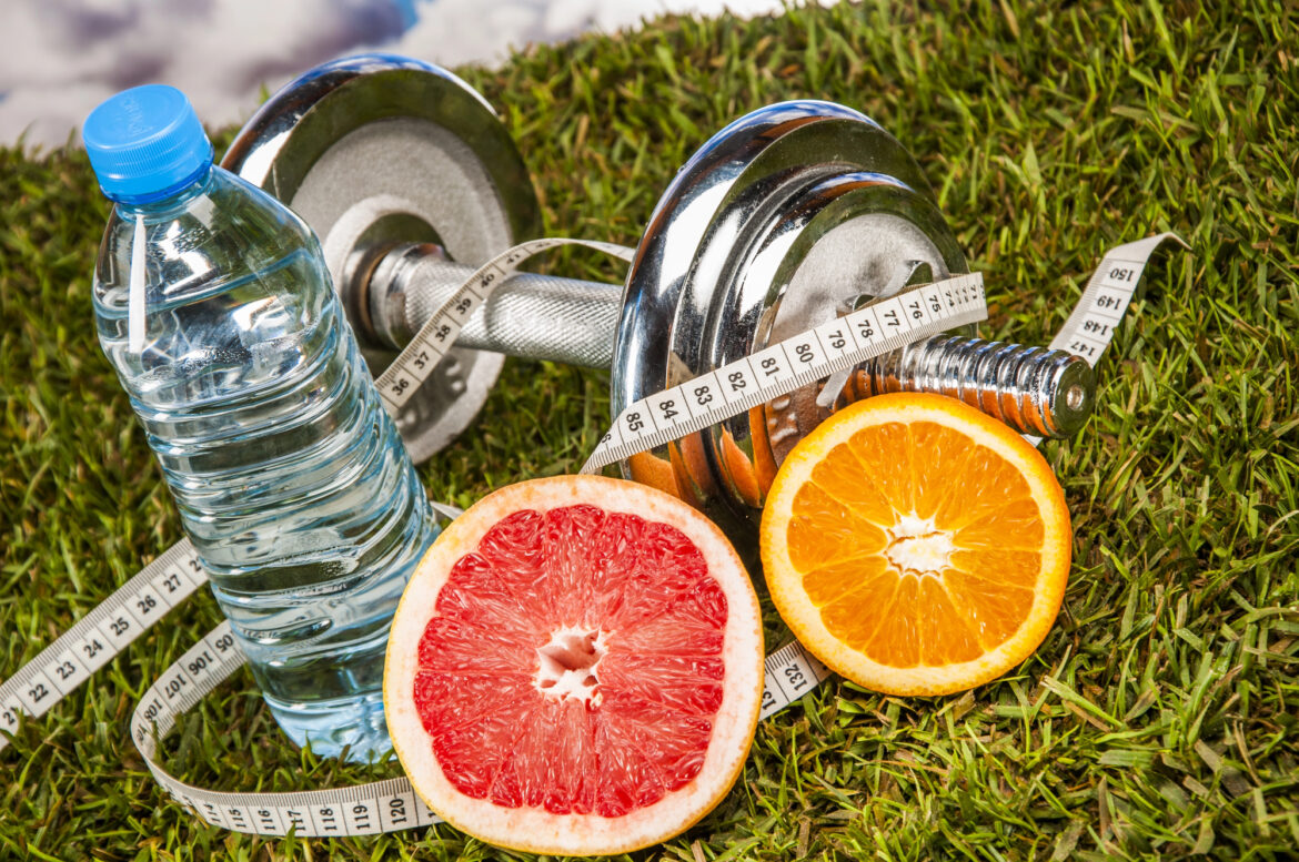 A water bottle, weight, measuring tape, orange, and grapefruit sit in the grass together.